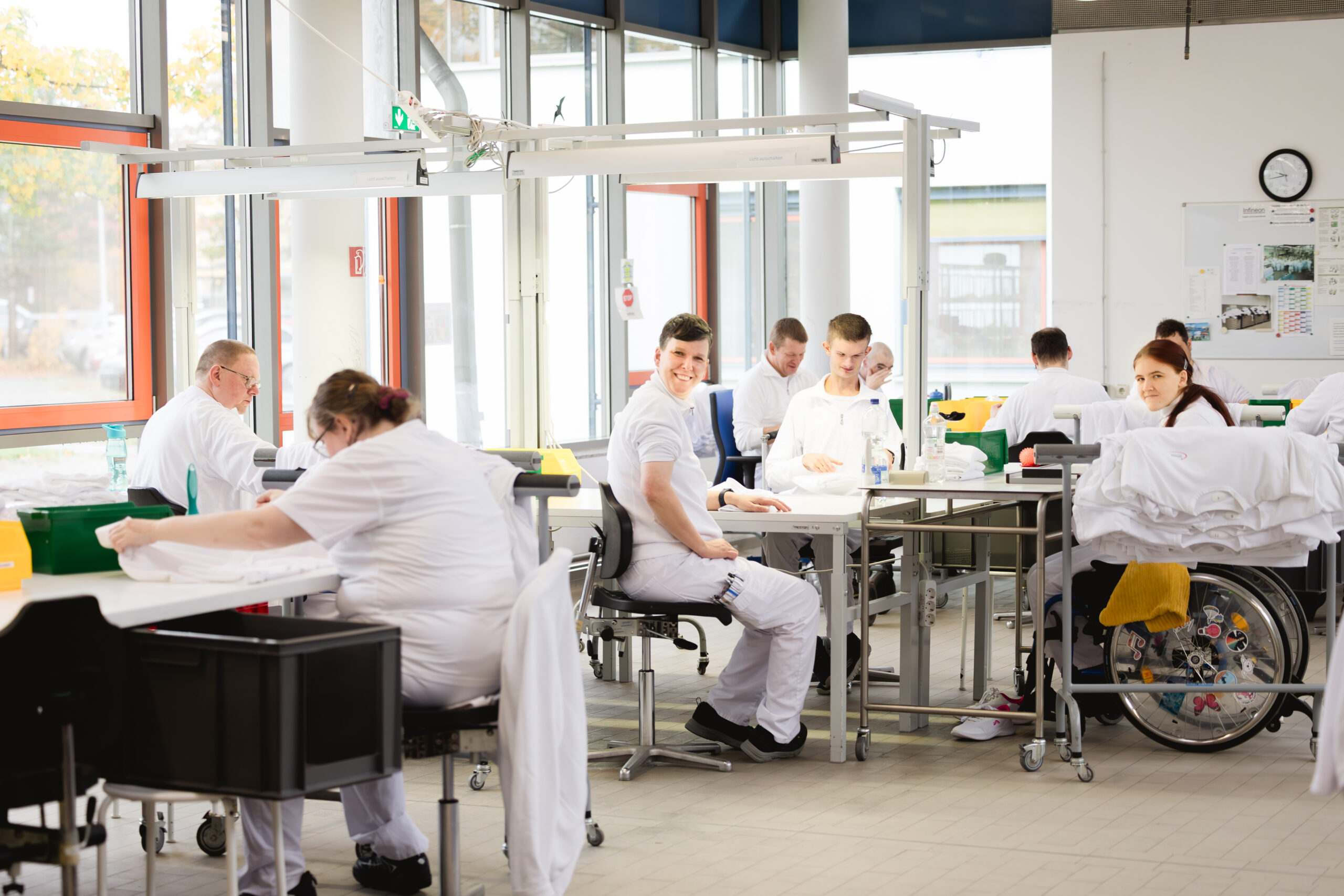 Mehrere Personen in weißen Arbeitskleidung arbeiten an Tischen in einem hellen, großen Arbeitsraum, eine Person sitzt im Rollstuhl.