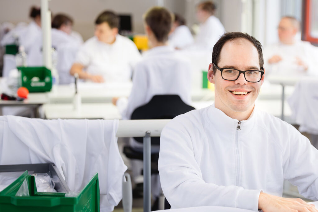 Ein Mann mit Brille und weißem Kittel sitzt lächelnd an einem Tisch in einer Wäscherei.