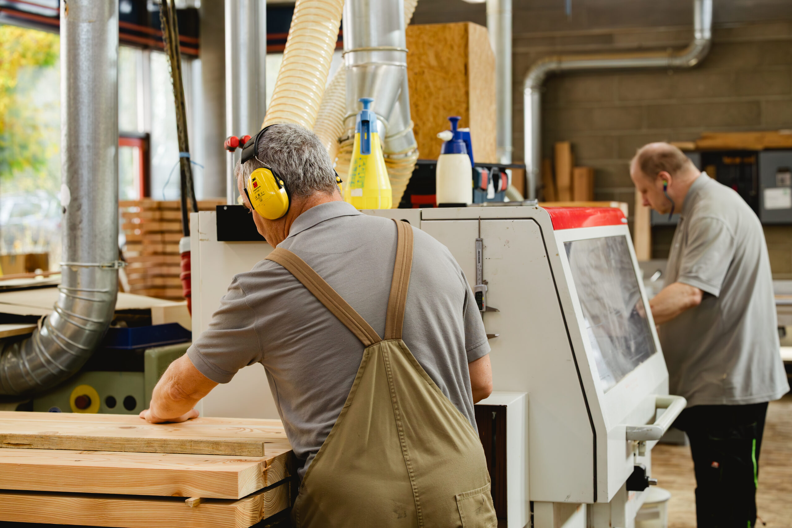 Zwei Männer mit Gehörschutz arbeiten an Maschinen in einer Schreinereihalle.
