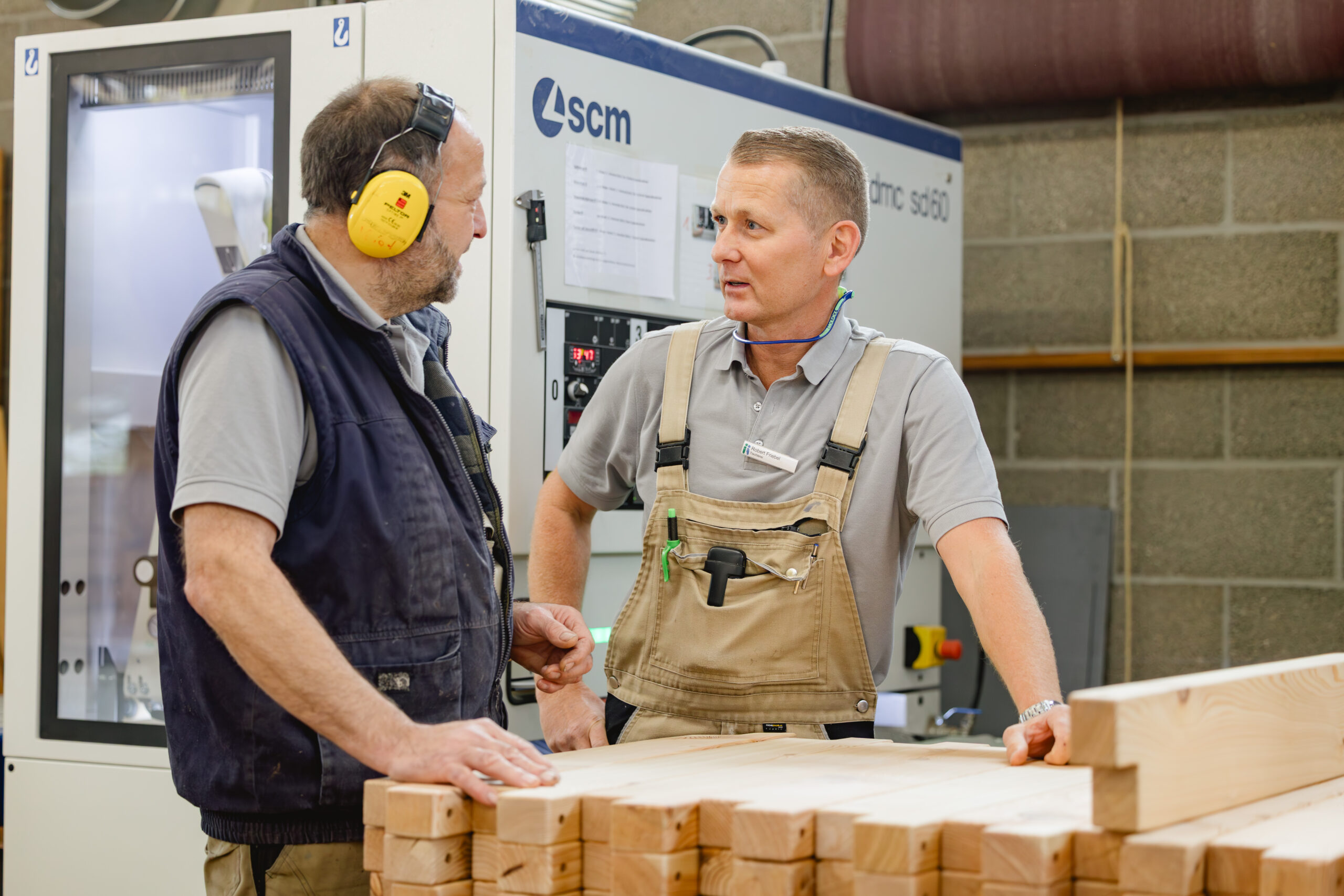 Zwei Männer sprechen miteinander an einem Holzstapel vor einer Industriemaschine.