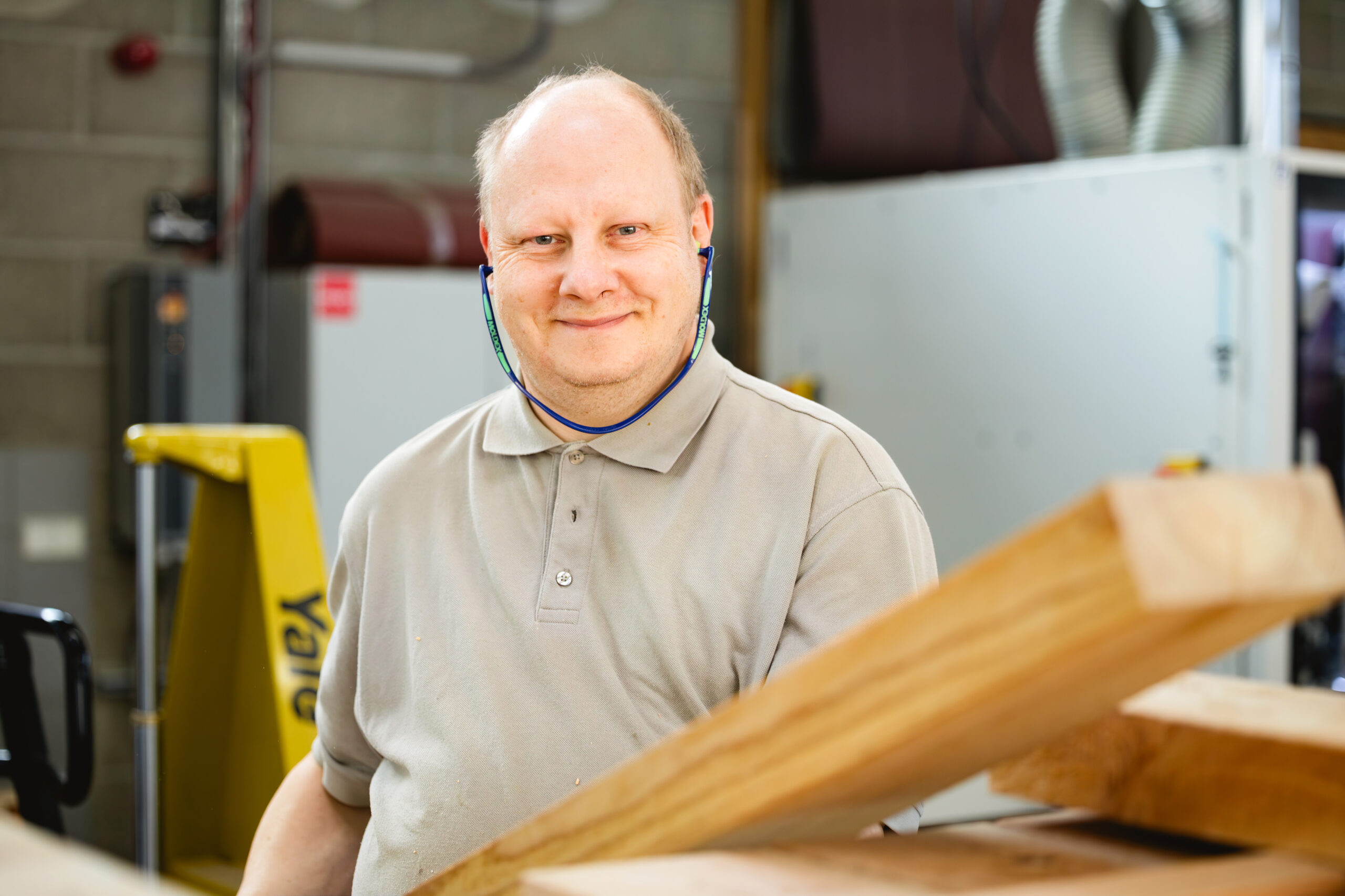 Ein Mann im grauen Poloshirt lächelt hinter einem Holzbrett in einer Schreinereihalle.