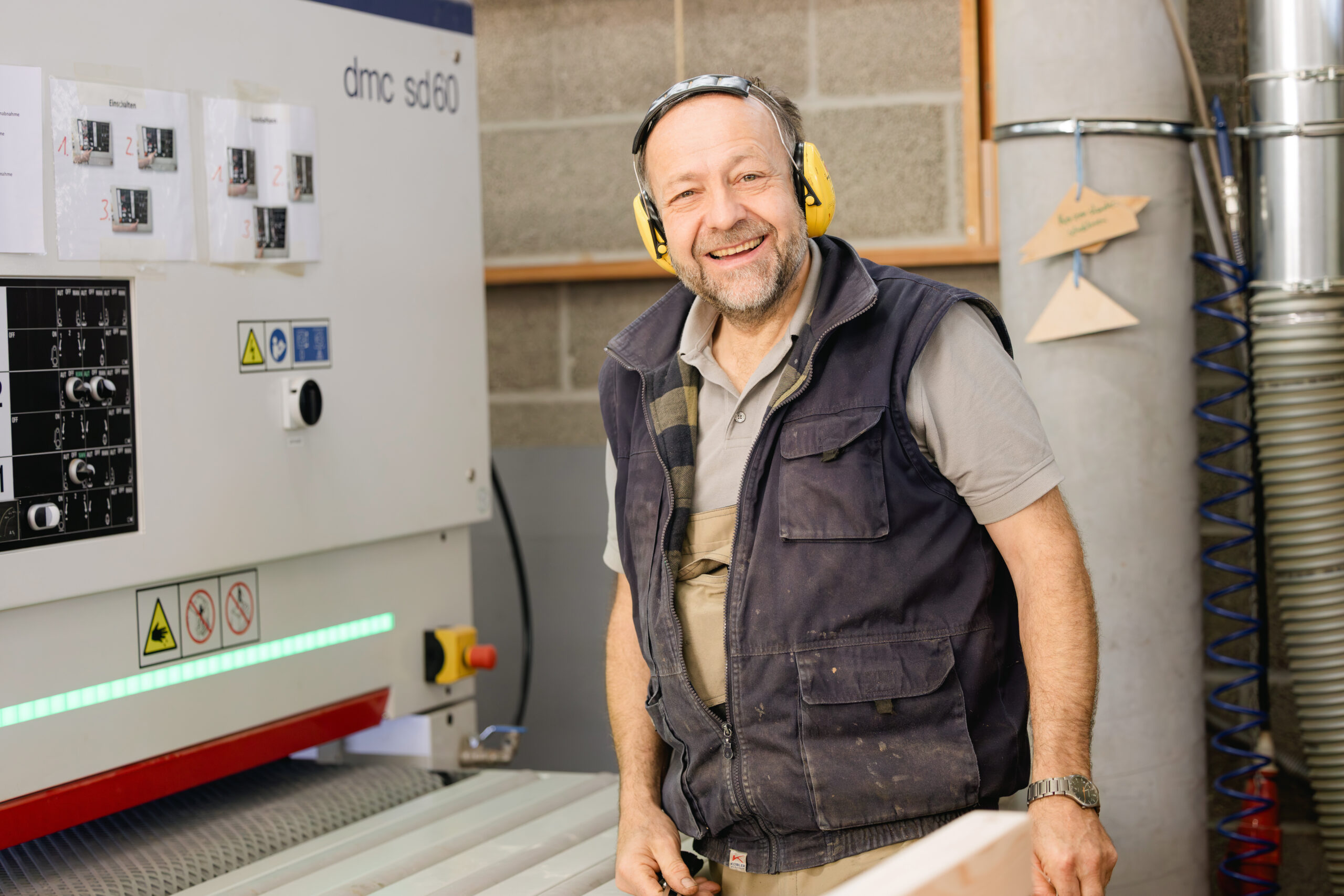 Ein Mann mit gelbem Gehörschutz und Schutzbrille lächelt neben einer Holzbearbeitungsmaschine.