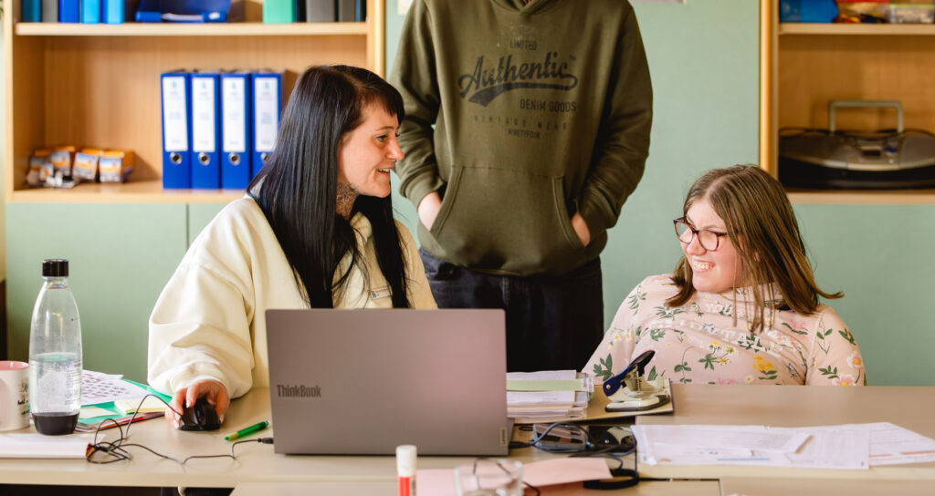 Zwei Frauen sitzen lachend an einem Schreibtisch mit Laptop, eine dritte Person steht im Hintergrund.