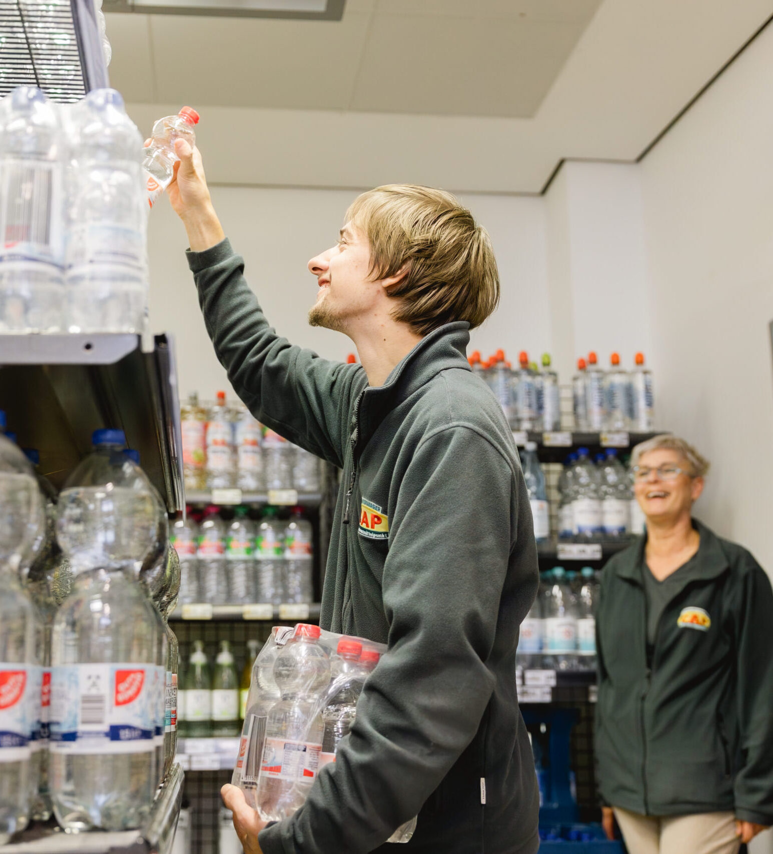 Ein junger Mann räumt Wasserflaschen ins Regal, eine Kollegin lacht im Hintergrund.