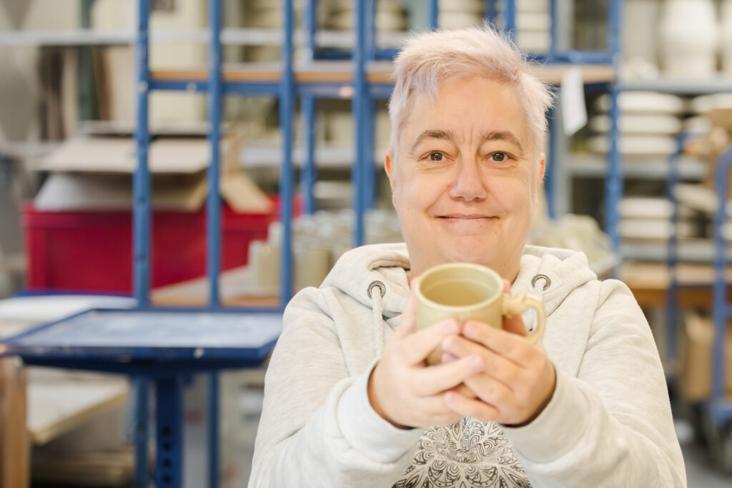 Eine Frau mit kurzem weißem Haar hält lächelnd eine kleine Keramiktasse in die Kamera, im Hintergrund Töpfereiregale.