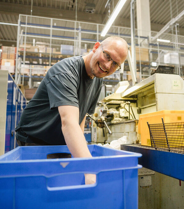 Ein Mann mit Brille lächelt und lehnt sich über eine blaue Transportkiste neben einer Industriemaschine in einer Produktionshalle.