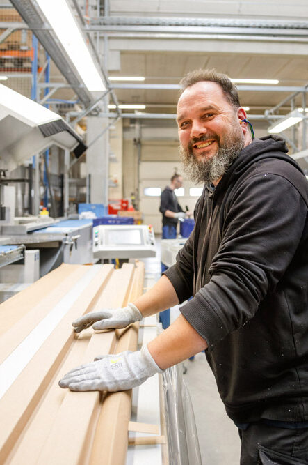 Ein bärtiger Mann mit Arbeitshandschuhen lächelt an einer Holzbearbeitungsmaschine in einer großen Produktionshalle in die Kamera.