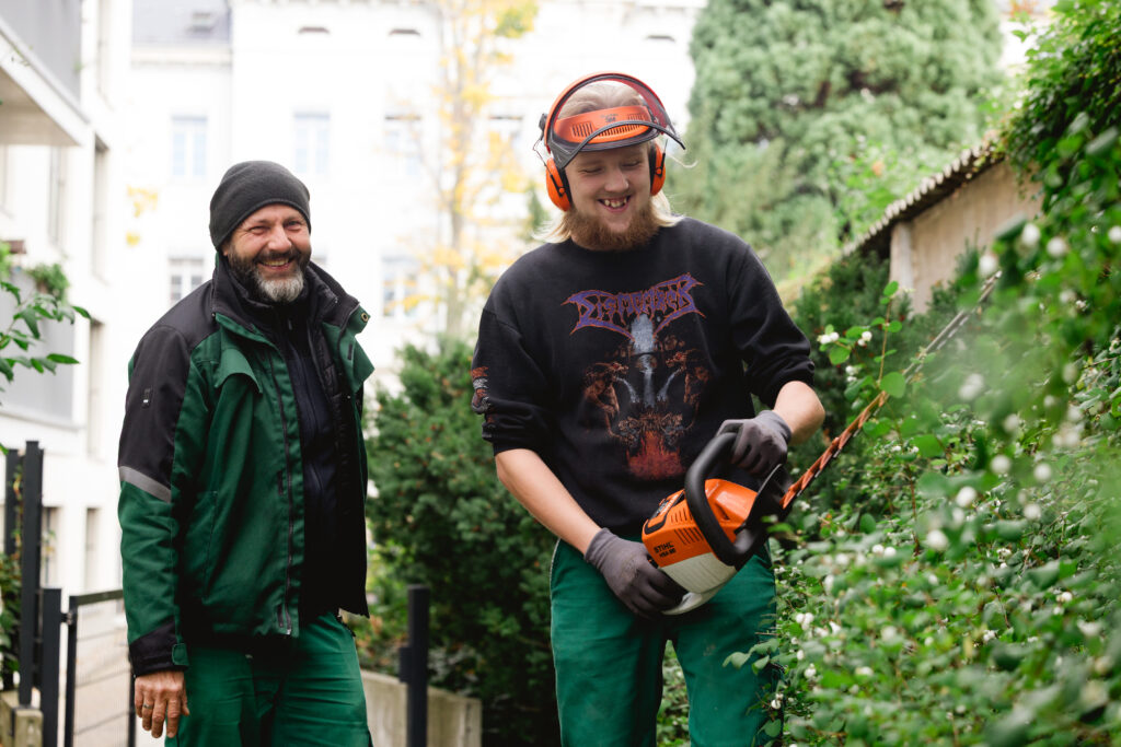 Zwei Männer in grüner Arbeitskleidung schneiden lachend eine Hecke mit einem Akku-Heckenschneider.