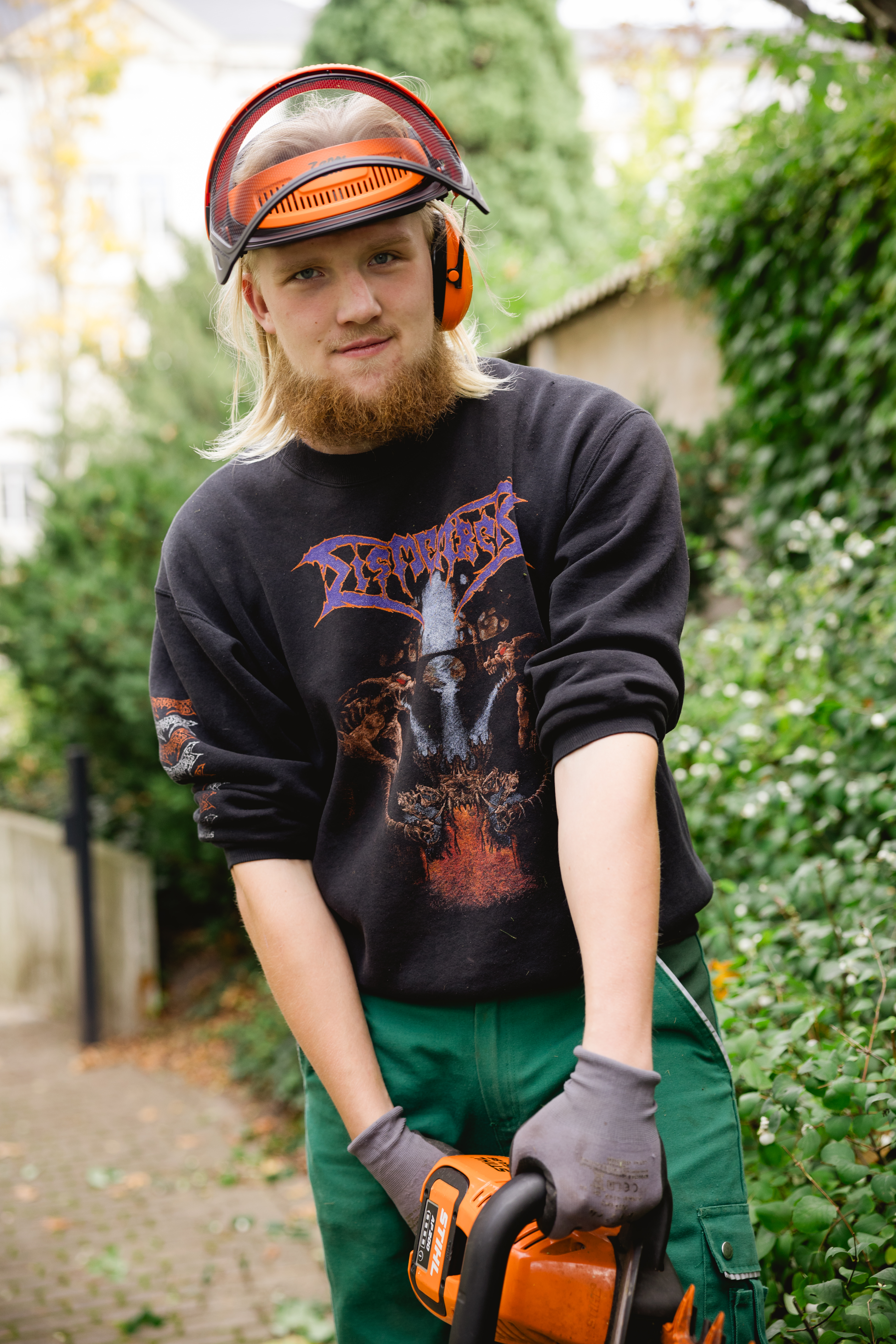Ein Mann mit Schutzhelm und Freischneider mäht eine Rasenfläche vor einem Wohngebäude.