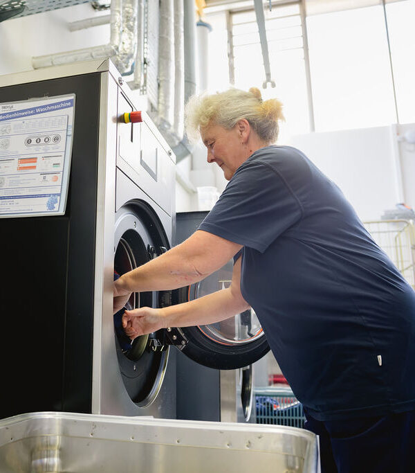 Eine Frau öffnet die Luke einer Industriewaschmaschine und lädt Wäsche ein.
