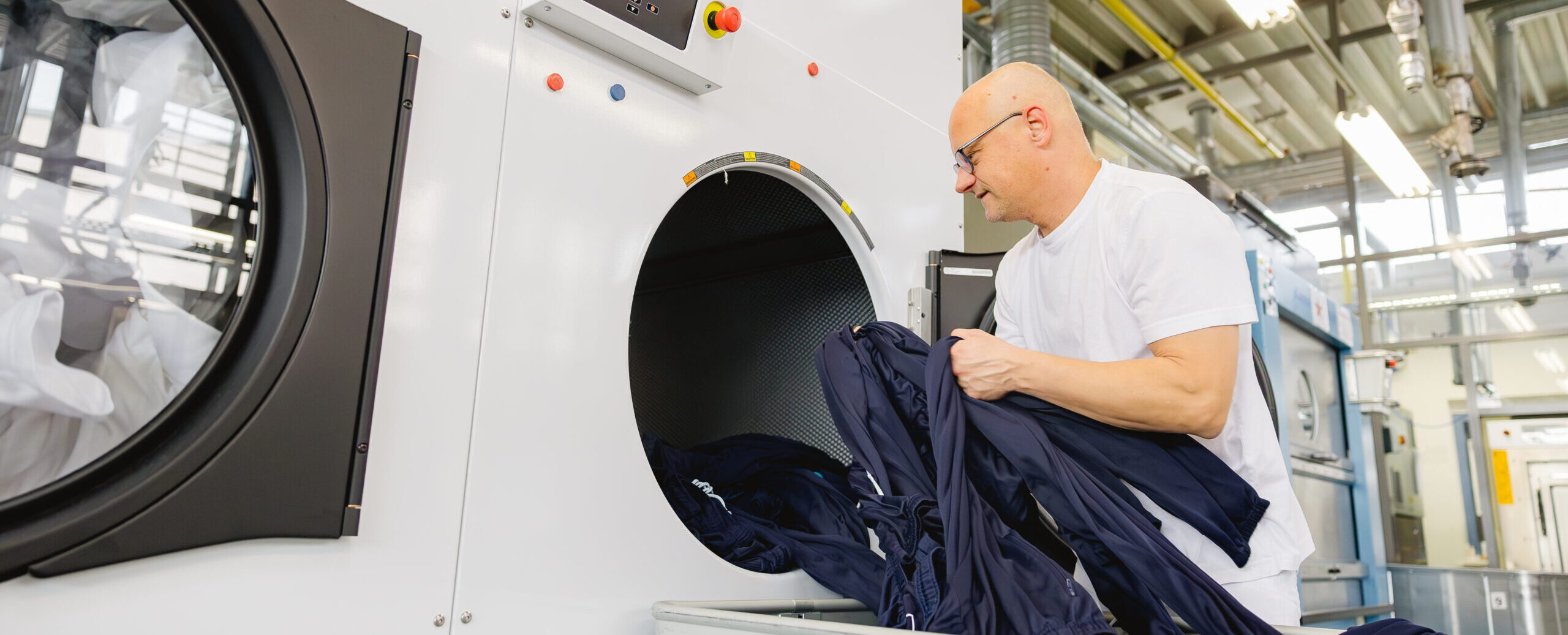 Ein Mann in weißem T-Shirt lädt dunkelblaue Wäsche in einen großen Industrietrockner.