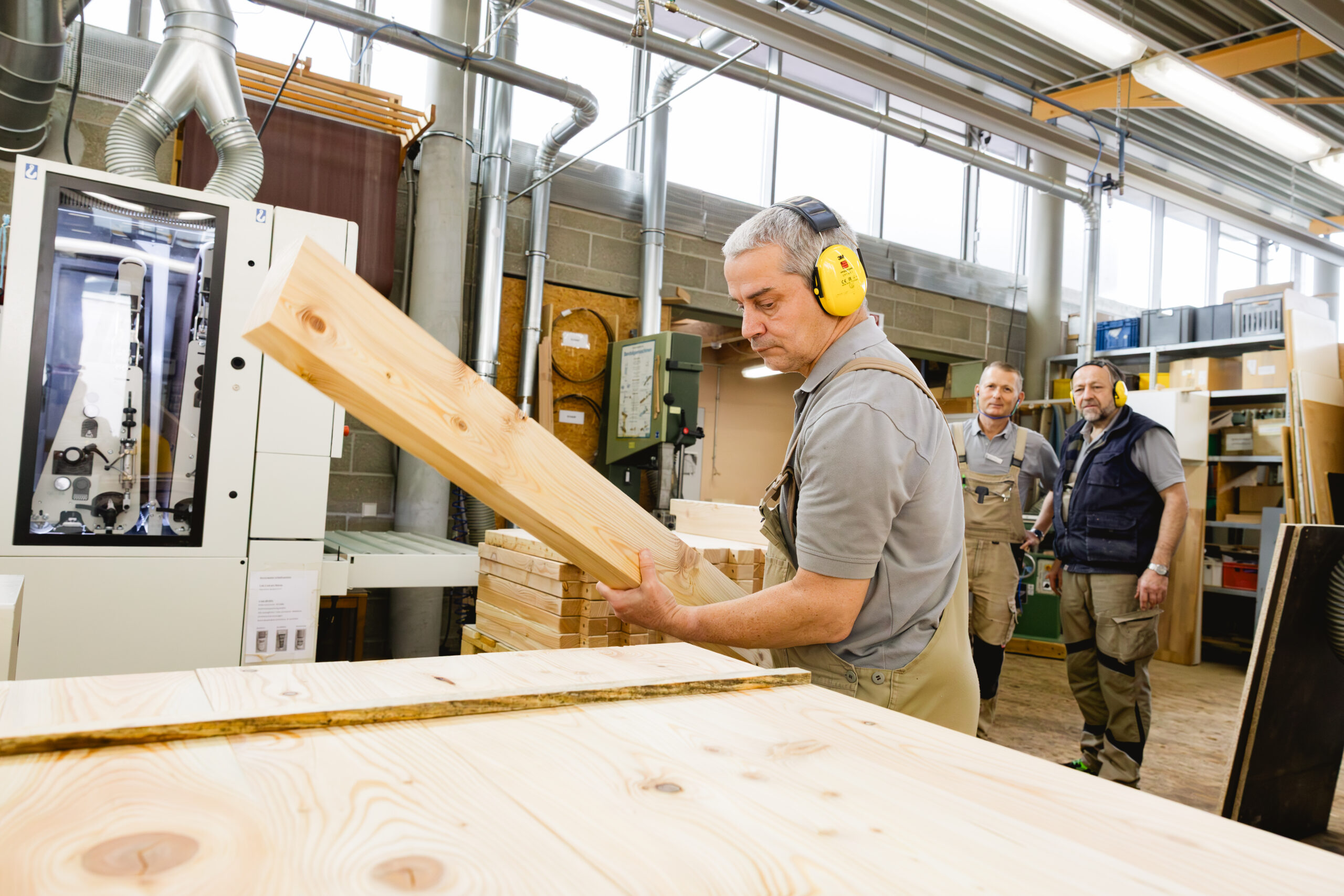 Ein Mann mit Gehörschutz hebt ein Holzbrett, zwei Kollegen beobachten ihn im Hintergrund.
