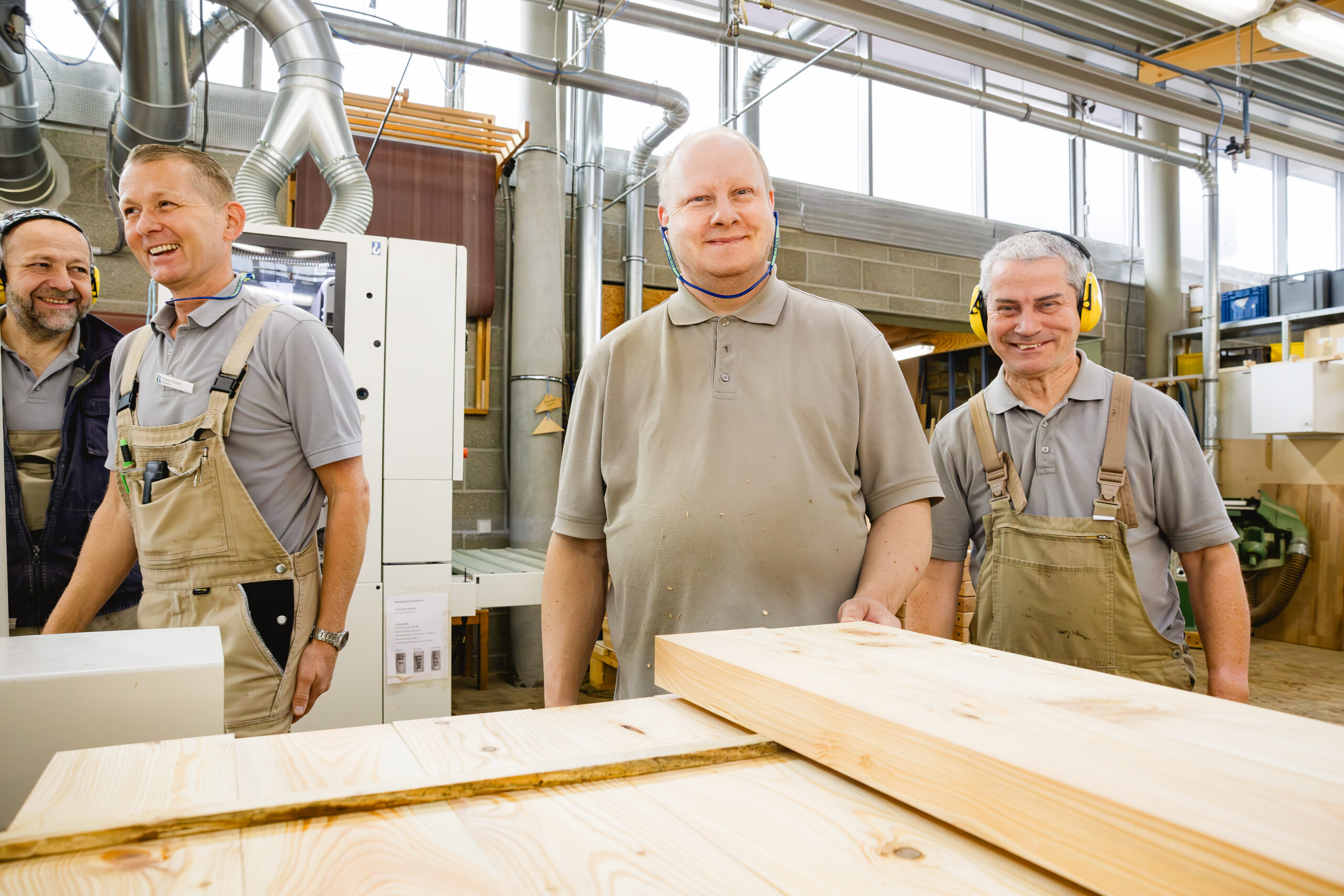 Vier Männer stehen lächelnd vor Holzbrettern und Maschinen in einer Schreinereihalle.