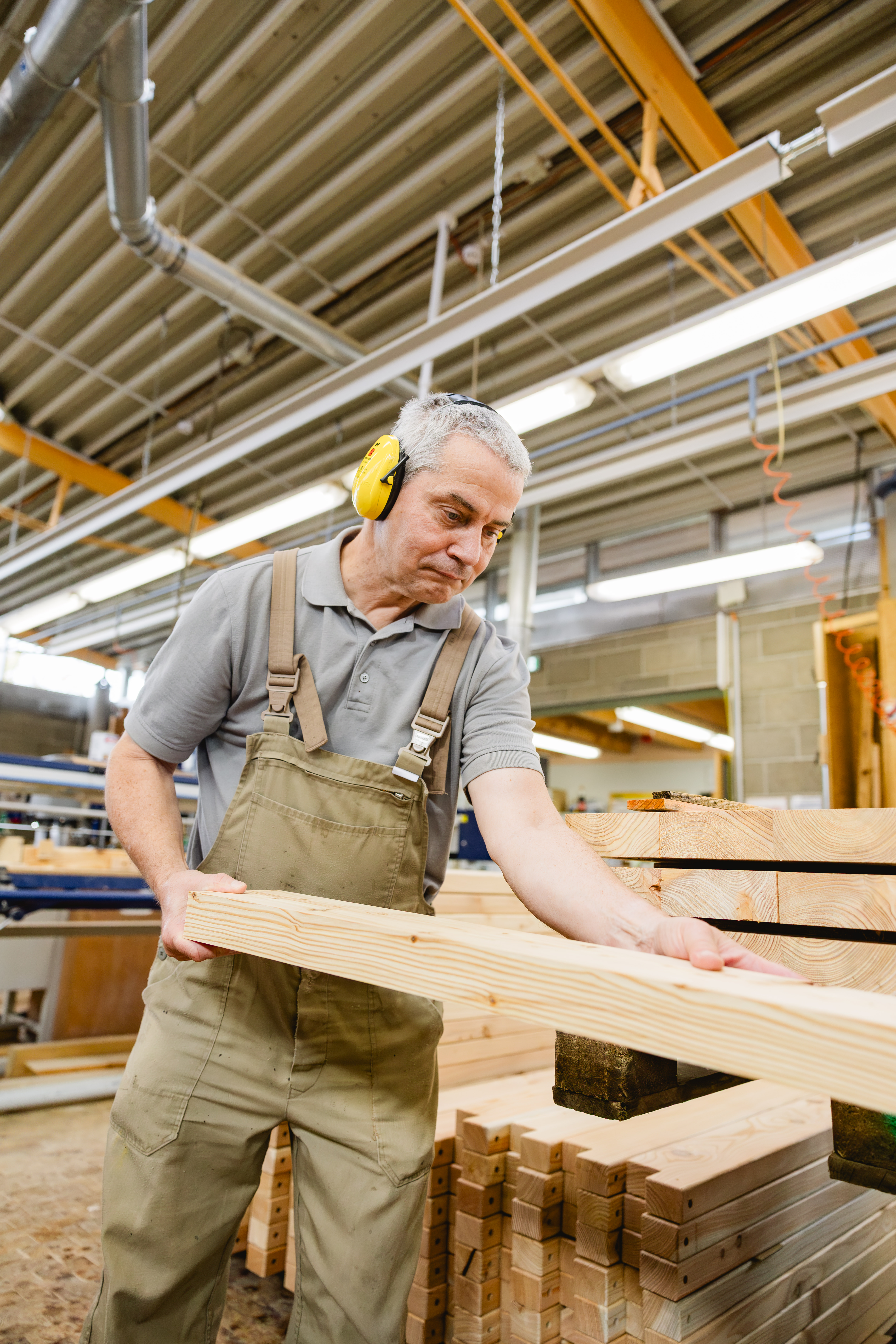 Ein Mann mit gelbem Gehörschutz hebt ein Holzbrett und prüft es in einer Schreinereihalle.