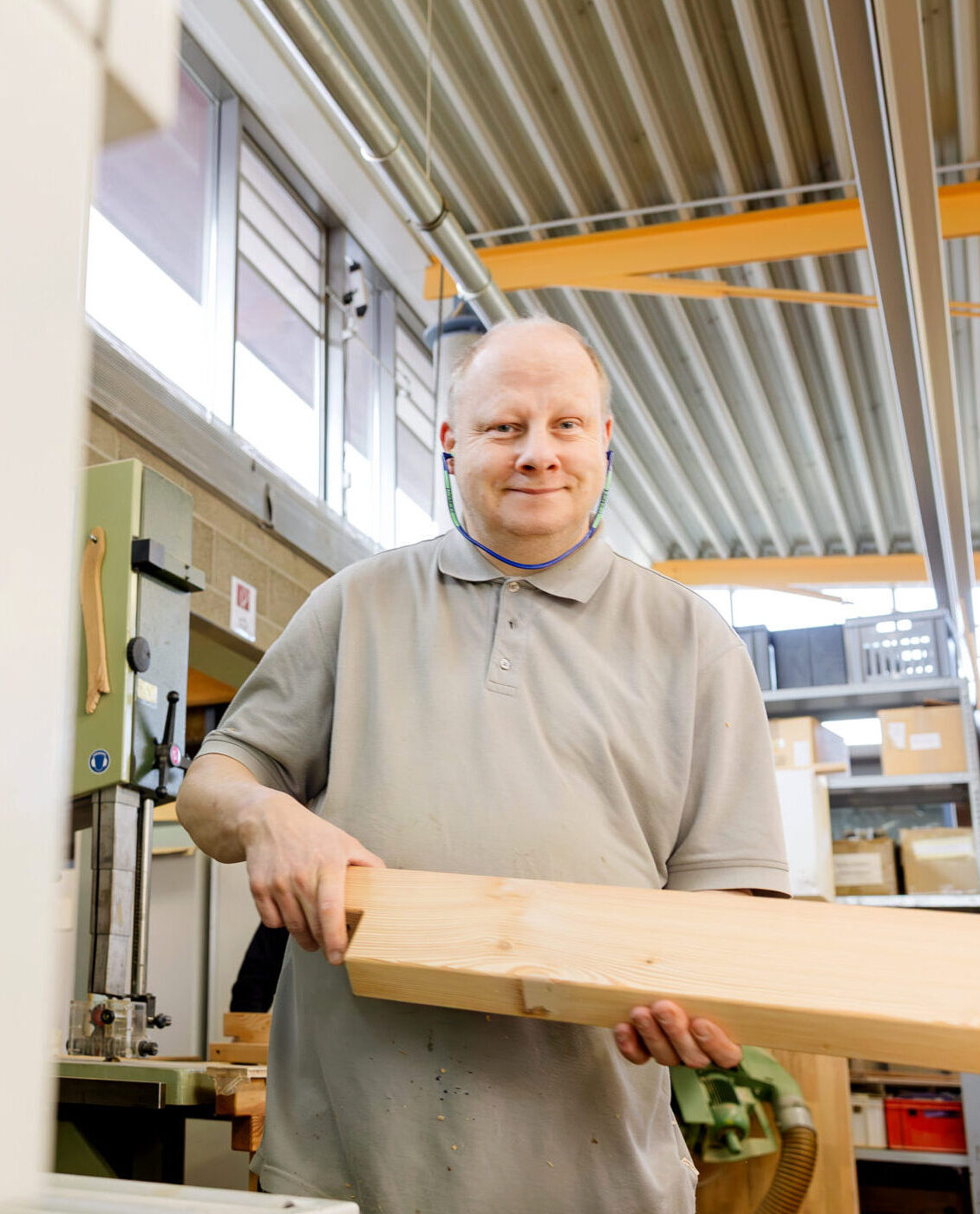 Ein Mann im grauen Poloshirt trägt lächelnd ein Holzbrett durch eine helle Schreinereihalle.