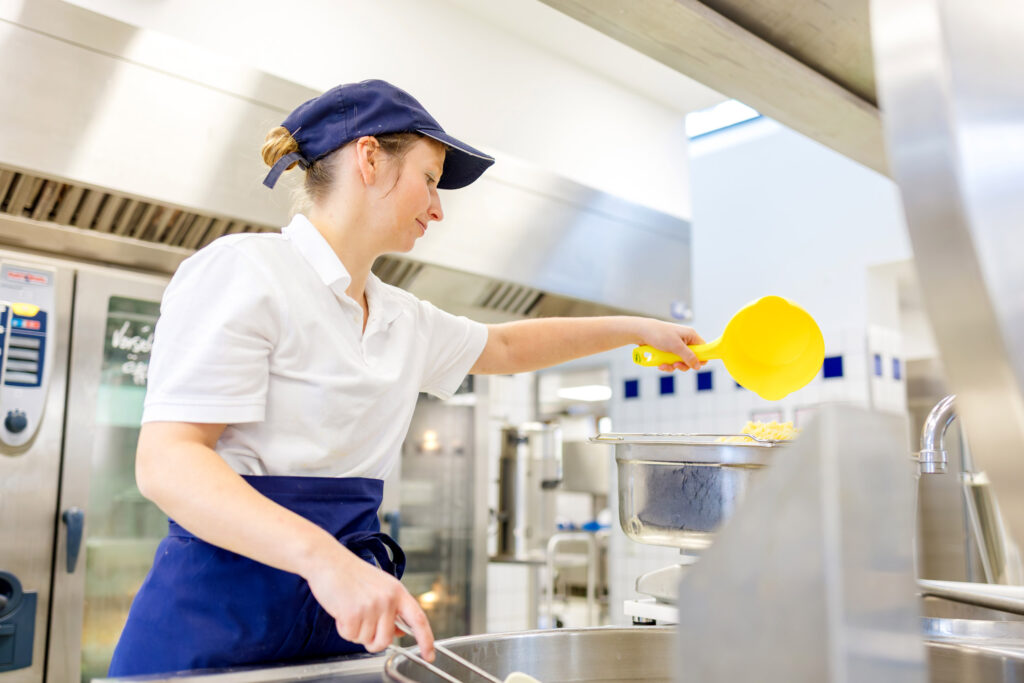 Eine Frau in weißem Poloshirt, blauer Schürze und Mütze portioniert Pommes in einer Großküche.