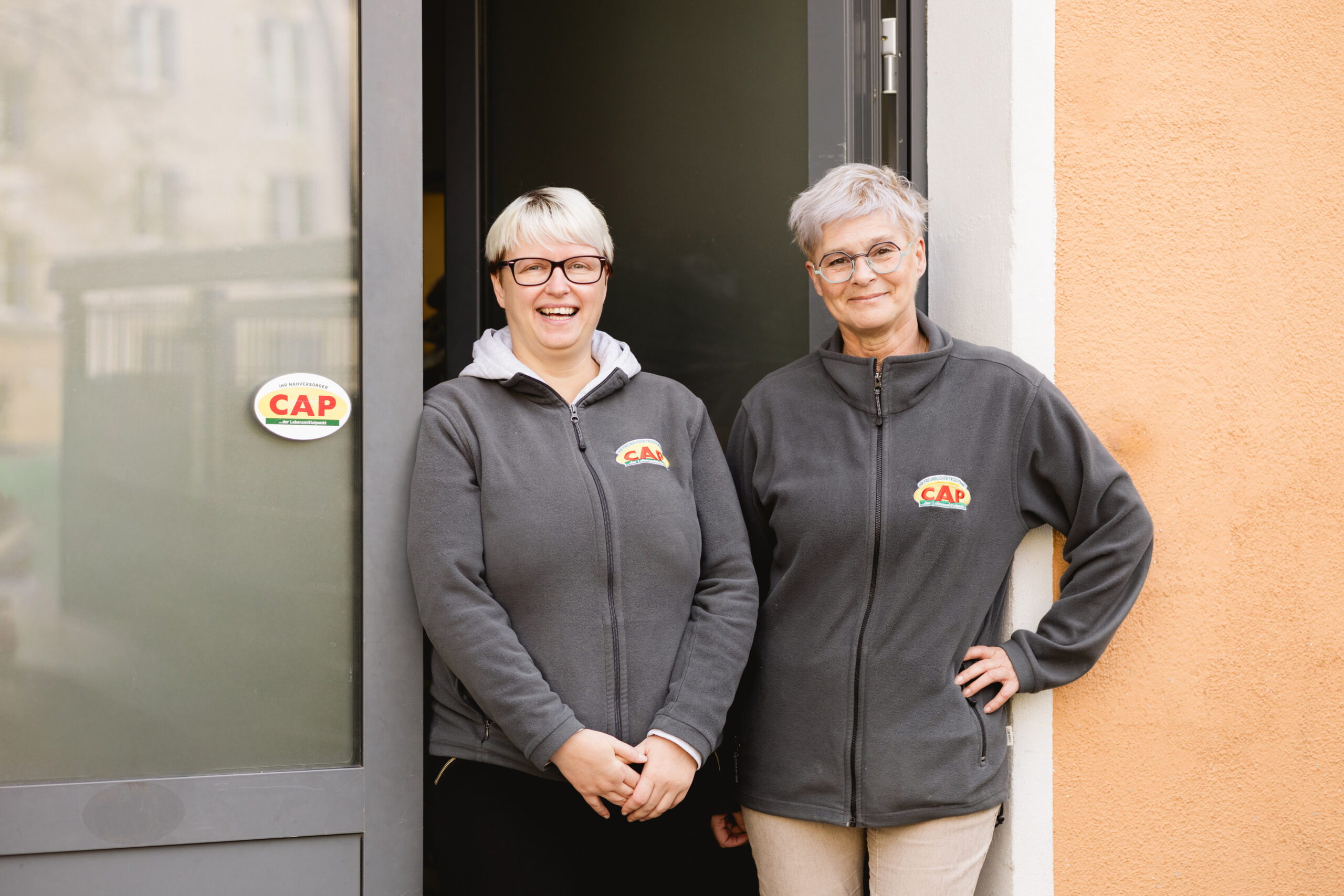 Zwei Frauen in grauen CAP-Fleecejacken stehen lächelnd vor der Eingangstür des Markts.