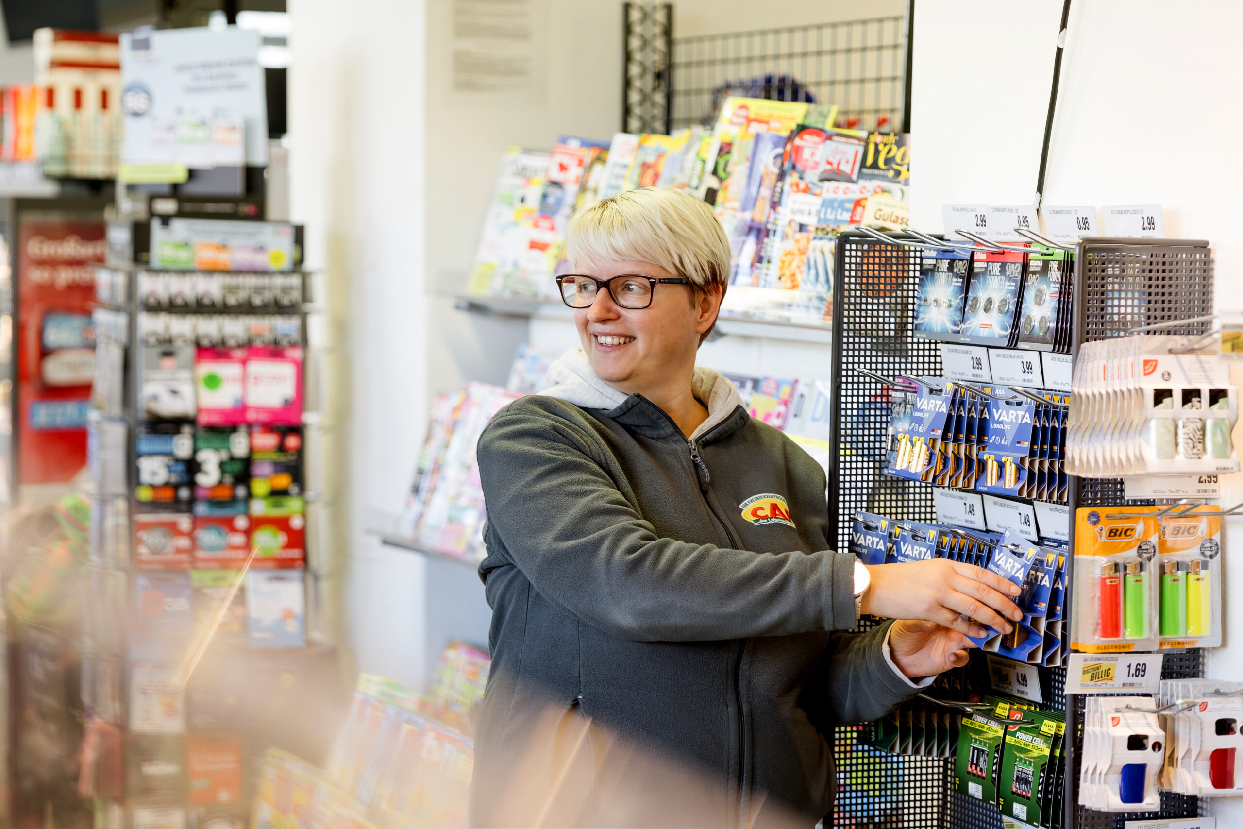 Eine Frau mit weißem, kurzen Haar befüllt lächelnd ein Batterieregal im Markt.