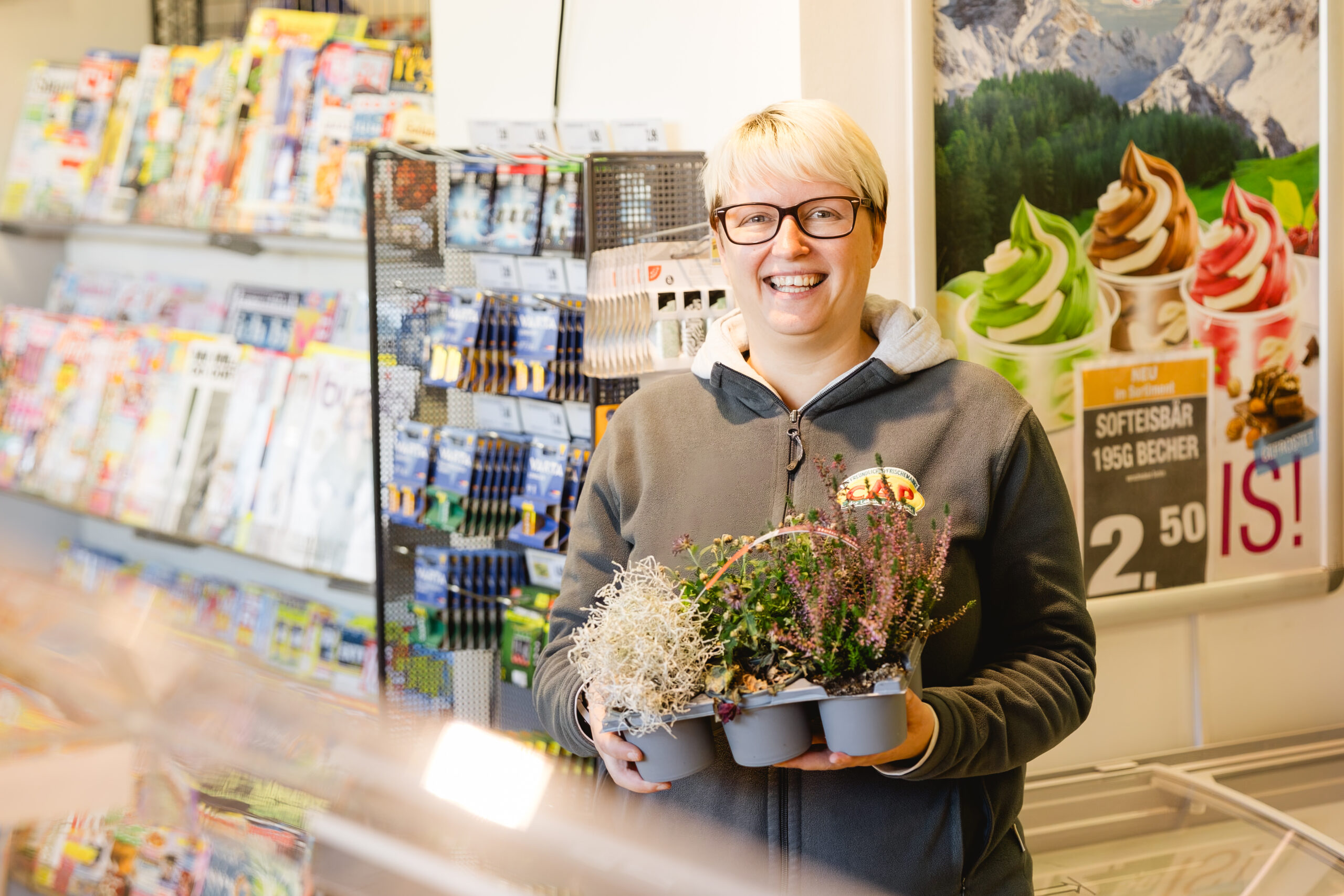 Eine Frau mit weißem Kurzhaar hält lächelnd einen Pflanzentopf mit Heidekraut im Markt.