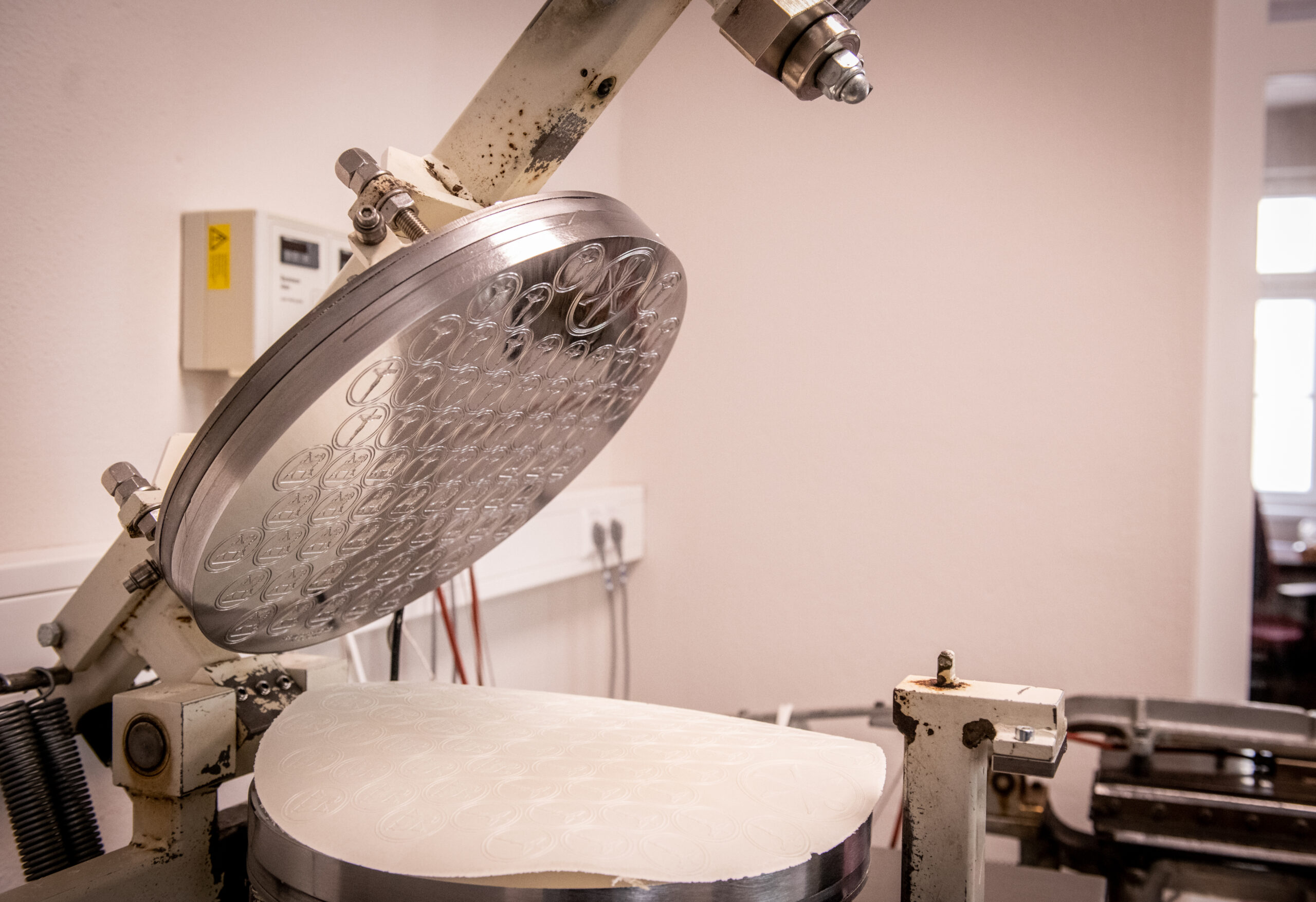 Eine runde Metallpresse mit Prägefläche öffnet sich über einem weißen Oblatenblatt auf einer Industriemaschine.