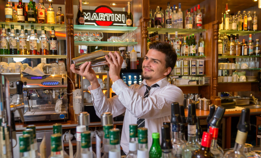 Ein junger Barkeeper in weißem Hemd schüttelt lächelnd einen Cocktailshaker hinter der voll bestückten Bar.