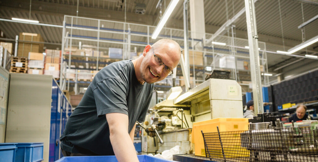 Ein Mann mit Brille lächelt und lehnt sich über eine blaue Transportkiste neben einer Industriemaschine in einer Produktionshalle.
