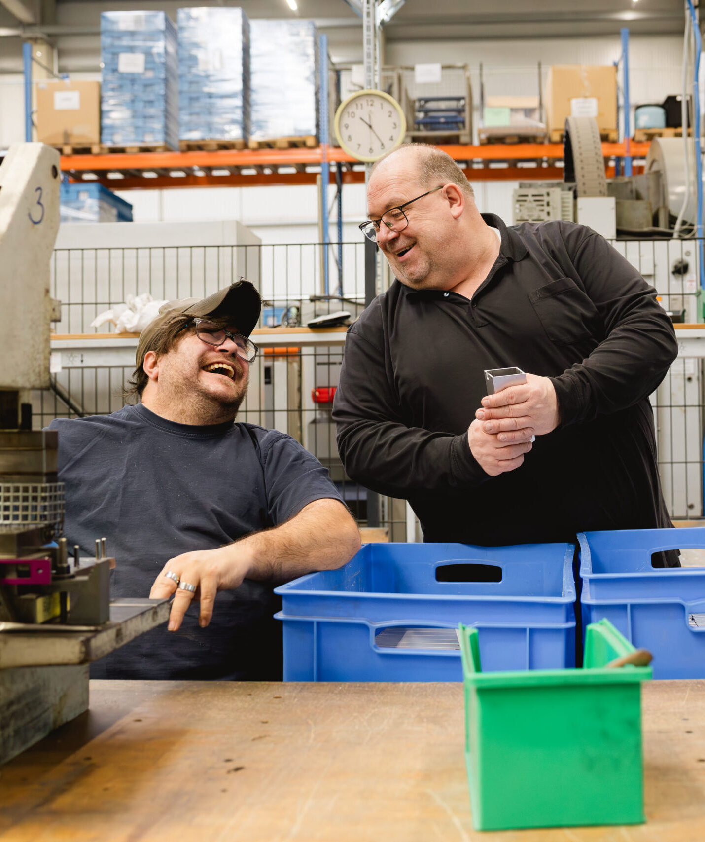 Zwei Personen lachen gemeinsam an einem Werktisch mit blauen Transportkisten in einer Produktionshalle.