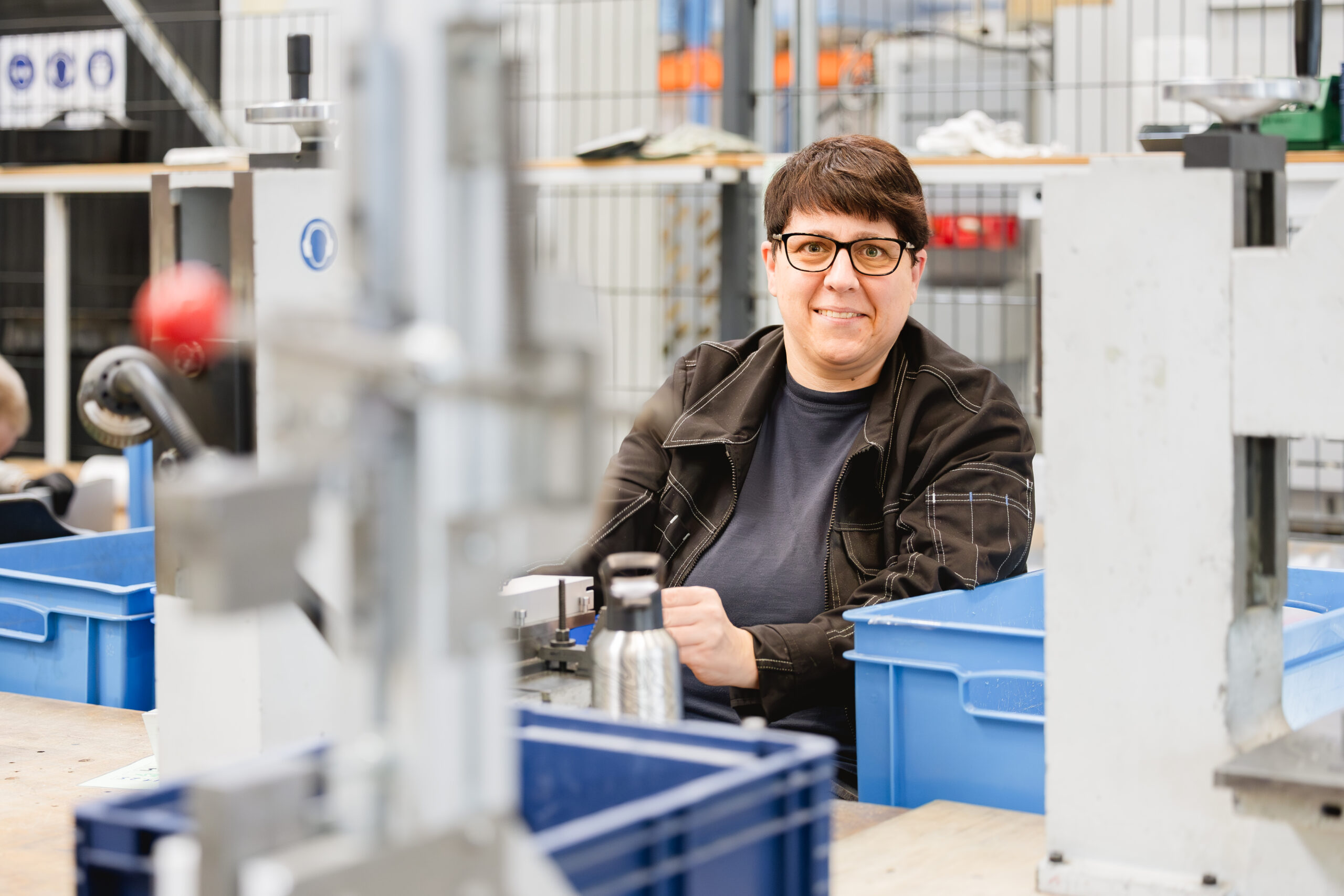 Eine Frau mit Brille und schwarzer Jacke lächelt zwischen Metallgestellen und blauen Transportkisten in einer Werkstatt.