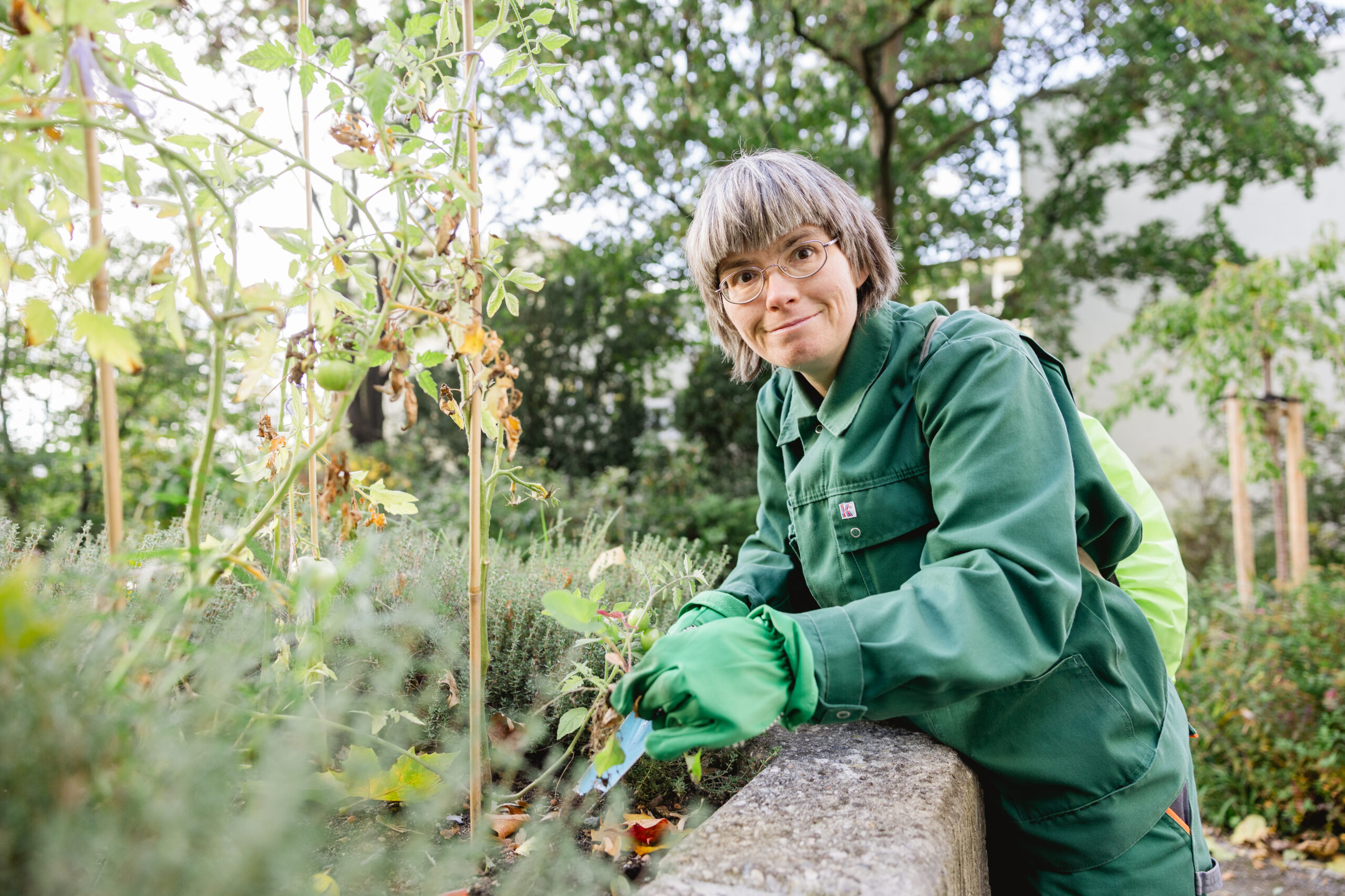 Portrait: Elfie. Eine lächelnde Frau mit grauem Bob und Brille, bei der Gartenarbeit.
