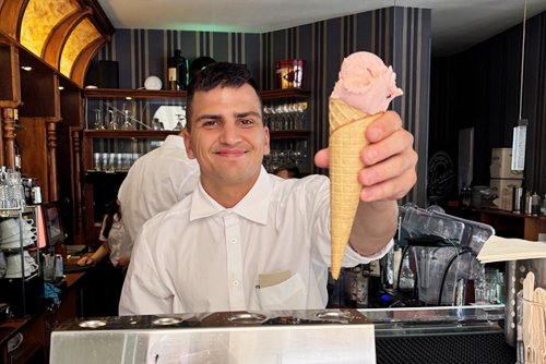 Portrait: Flavio. Ein lächelnder Mann mit braunen Haaren serviert Eiscreme.