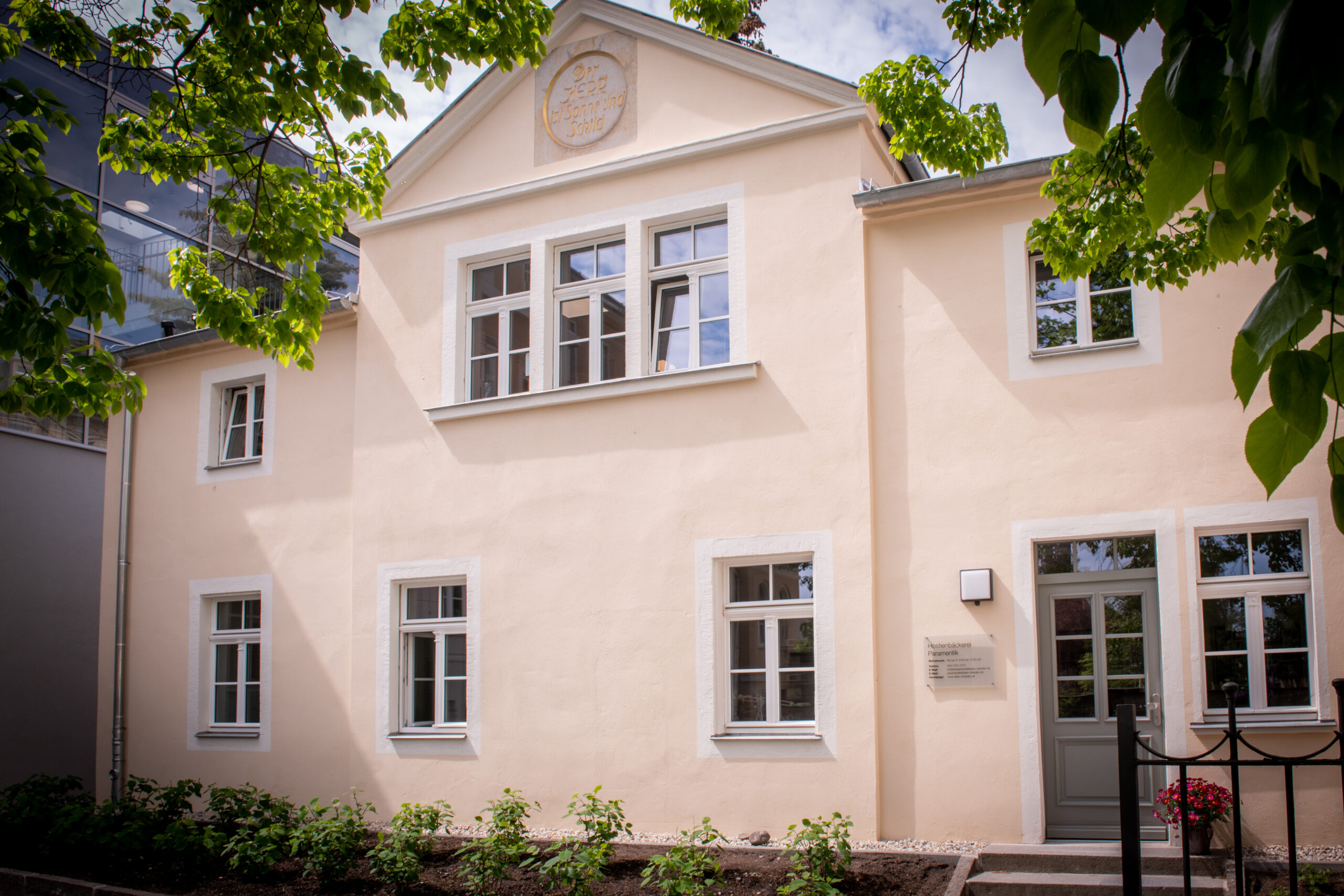 Cremefarbe Fassade der Hostienbäckerei mit Giebel und goldenem Medaillon, umgeben von Bäumen.